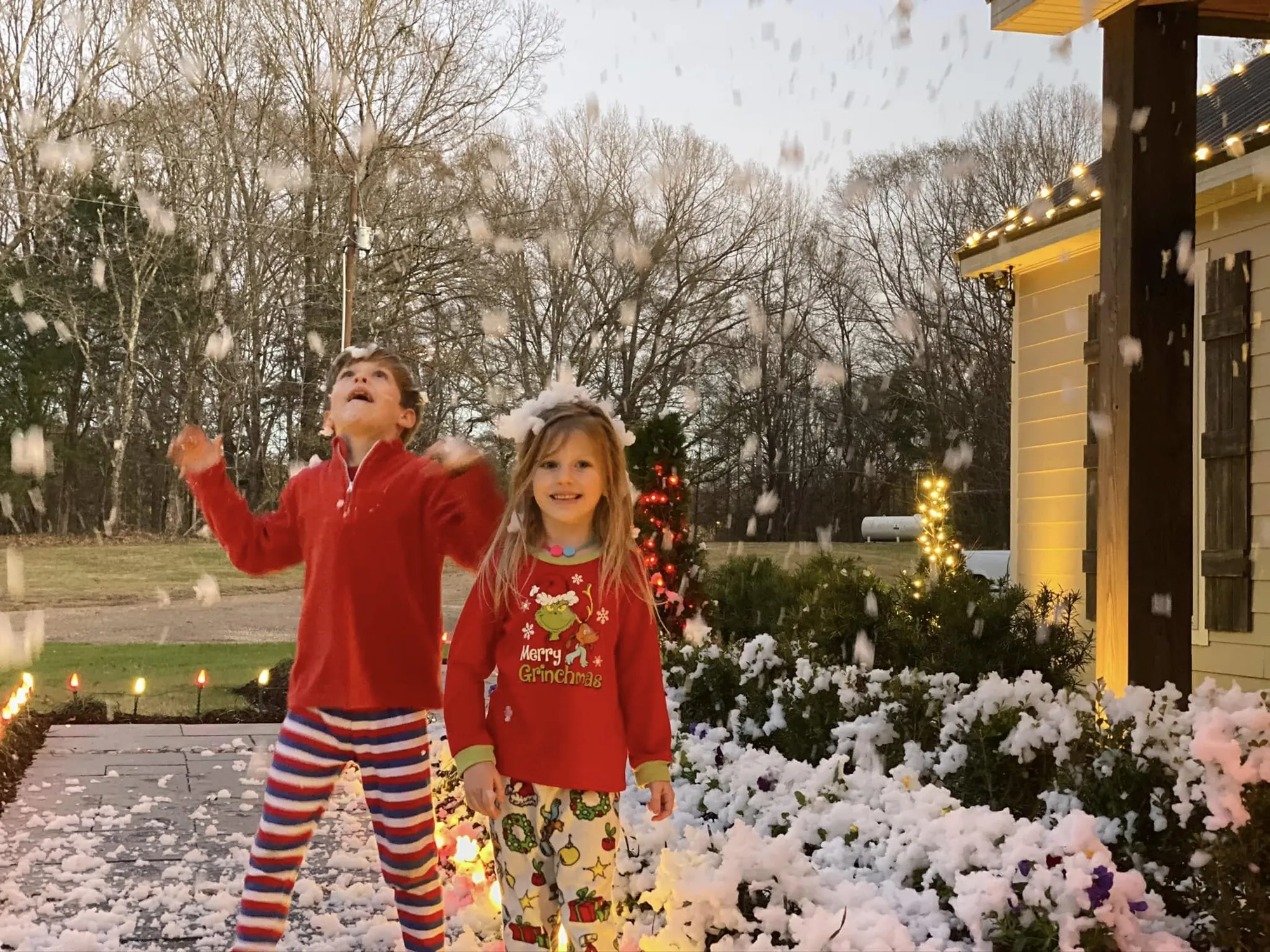 Two children in holiday pajamas playing in fake snow beside a lit home